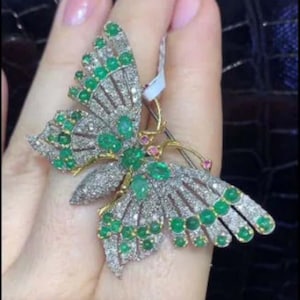 Puede incluir: Un broche de mariposa de oro con piedras preciosas de color verde esmeralda y transparentes. La mariposa tiene alas detalladas y un cuerpo delicado.