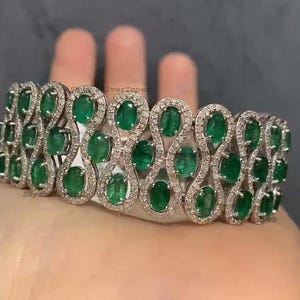 Puede incluir: Una pulsera de plata con esmeraldas ovaladas y pequeñas piedras claras. Las esmeraldas son de un verde vibrante, dispuestas en un patrón repetitivo. La pulsera se muestra en una mano.