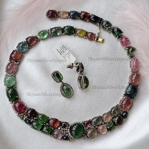 May include: A multi-colored gemstone necklace, bracelet, and earring set. The jewelry features a variety of oval and rectangular gemstones in shades of green, pink, and brown, set in a silver metal. The earrings have a dangling design.