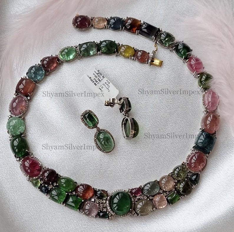 May include: A multi-colored gemstone necklace, bracelet, and earring set. The jewelry features a variety of oval and rectangular gemstones in shades of green, pink, and brown, set in a silver metal. The earrings have a dangling design.