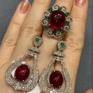 Könnte beinhalten: Ein Schmuckset mit einem großen Ring mit rotem Edelstein und passenden Ohrringen. Der Ring hat einen großen ovalen roten Stein, der von kleinen grünen und klaren Steinen umgeben ist. Die Ohrringe haben ein tropfenförmiges Design mit einem roten Stein.
