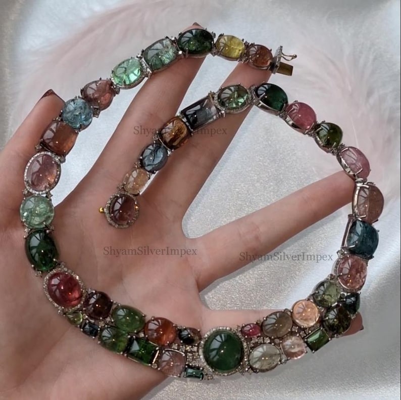 May include: A multi-colored gemstone necklace featuring oval and round-cut stones in shades of green, pink, blue, and brown. The necklace is set in a silver metal with a clasp closure. The gemstones are surrounded by small, clear stones.