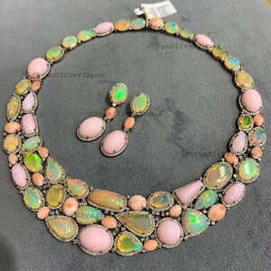 May include: A necklace and earring set featuring a variety of gemstones in shades of pink, orange, and green. The necklace and earrings are set in a silver metal with small, clear stones around the edges. The gemstones are oval, round, and irregular shapes.