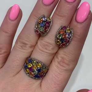 Può includere: Un set di gioielli con un anello e orecchini abbinati. L'anello è su un dito e gli orecchini sono accanto. Ogni pezzo è incrostato con una varietà di pietre preziose colorate, tra cui blu, giallo, rosa e verde, incastonate in un metallo argentato.