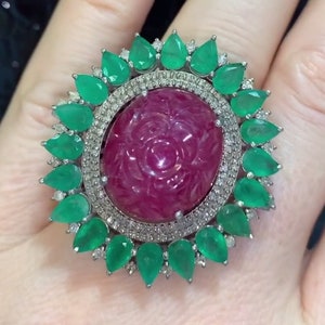 Op de afbeelding: Een grote zilveren ring met een gegraveerde robijn in het midden, omringd door een halo van kleinere diamanten en smaragdgroene stenen.