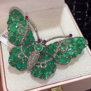 May include: A silver butterfly brooch with green emeralds and small diamonds. The butterfly has a detailed design with a black outline.