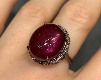 May include: A striking ruby ring featuring a large, oval-cut gemstone. The deep red ruby is set in a dark metal band, encircled by a halo of small, sparkling stones. The ring is a bold statement piece, highlighting the ruby's rich colour and brilliance.
