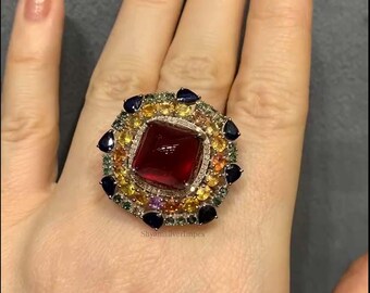 Könnte beinhalten: Ein auffälliger Ring mit einem quadratischen, tiefroten Edelstein im Zentrum, umgeben von einem Kranz aus kleineren, farbigen Edelsteinen in Gelb-, Blau-, Grün- und Orangetönen. Der Ring ist auf einem silbernen Band gefasst und wird auf einer Hand präsentiert.