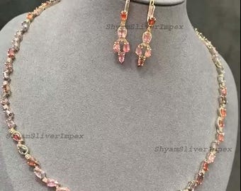 May include: A rose gold necklace and matching earrings set. The necklace and earrings feature a series of oval-shaped, multi-colored gemstones in shades of pink, peach, and clear. The jewelry is displayed on a gray mannequin.