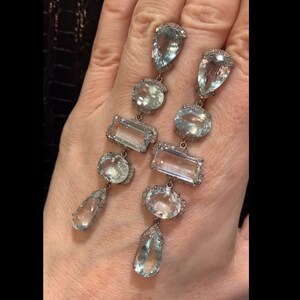 May include: A pair of long, silver dangle earrings with multiple aquamarine gemstones and diamond accents. The earrings feature a teardrop-shaped gemstone at the bottom, followed by a rectangular gemstone, a round gemstone, and a teardrop-shaped gemstone at the top.
