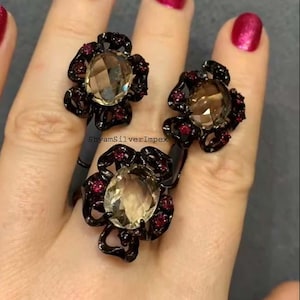 以下が含まれることがあります： 黒い金属仕上げの、花の形をした3つの指輪。各指輪は、中央に大きなファセットカットの薄茶色の宝石を特徴とし、その周りを小さな赤い宝石が囲んでいます。赤いマニキュアを塗った手の上に指輪が展示されています。