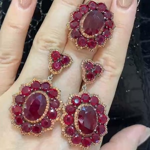 Puede incluir: Un par de pendientes colgantes con una gran piedra preciosa roja rodeada de piedras preciosas rojas más pequeñas. Los pendientes son de plata y tienen un diseño en forma de corazón. Los pendientes también vienen acompañados de un anillo con una gran piedra preciosa roja rodeada de piedras preciosas rojas más pequeñas.