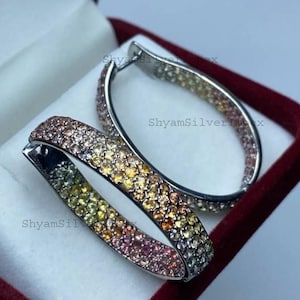 Könnte beinhalten: Ein Paar ovale Creolen mit einem Pavé-Besatz aus mehrfarbigen Edelsteinen in einem Farbverlauf von Gelb, Orange, Pink und Violett. Die Ohrringe sind aus silberfarbenem Metall gefertigt und tragen den Schriftzug ShyamSilverImpex.
