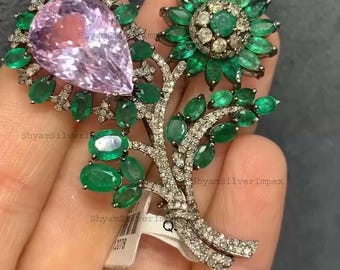 May include: A floral-themed brooch featuring a large, pear-shaped pink gemstone surrounded by green emeralds and small diamonds. The brooch has a tree-like design with additional emerald accents.
