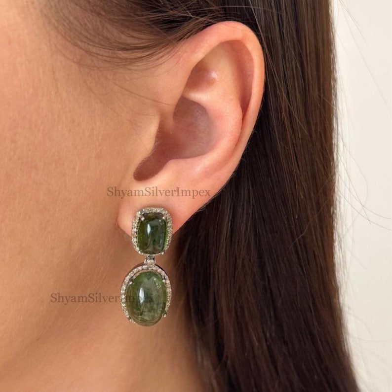 May include: Dangle earrings featuring two green gemstones, one square and one oval, set in a silver frame with a halo of small, clear stones. The earrings are displayed near an ear.