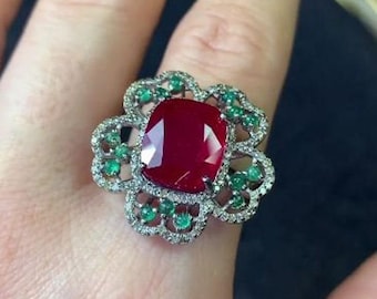 May include: A ring featuring a large, red gemstone surrounded by smaller green gemstones and diamonds. The ring is set in a silver band and has a floral design.