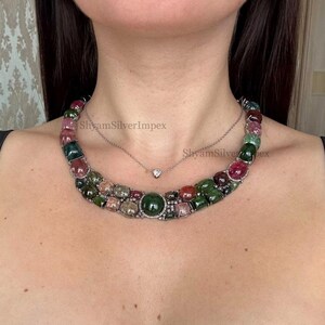May include: A multi-colored gemstone necklace with a variety of green, pink, and red stones. The necklace is layered with a delicate silver chain featuring a small diamond pendant. The necklace is a statement piece.