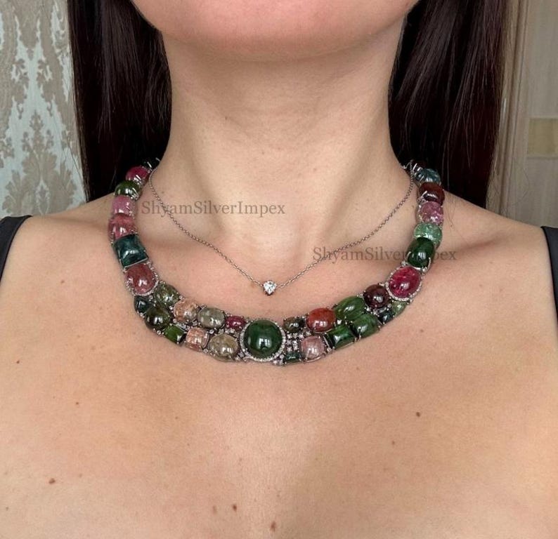 May include: A multi-colored gemstone necklace with a variety of green, pink, and red stones. The necklace is layered with a delicate silver chain featuring a small diamond pendant. The necklace is a statement piece.