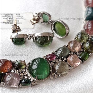 May include: A jewelry set featuring a necklace and earrings with large, colorful gemstones. The necklace and earrings have a silver setting. The gemstones are various colors, including green, pink, and clear. The brand name "ShyamSilverImpex" is visible.