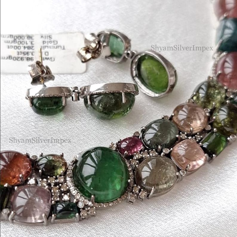 May include: A jewelry set featuring a necklace and earrings with large, colorful gemstones. The necklace and earrings have a silver setting. The gemstones are various colors, including green, pink, and clear. The brand name "ShyamSilverImpex" is visible.