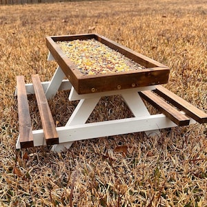 Könnte beinhalten: Ein weißer Holz-Picknicktisch mit einer braunen Holzplatte, die mit Vogelfutter gefüllt ist. Der Tisch steht auf trockenem Gras.