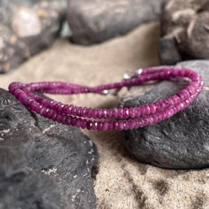 Könnte beinhalten: Ein zweireihiges Armband aus kleinen, facettierten, tief magentafarbenen Edelsteinperlen. Das Armband verfügt über einen metallischen, silberfarbenen Verschlussmechanismus, der die beiden Stränge verbindet und ein durchgehendes Design bildet.