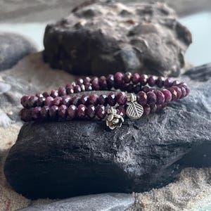 Könnte beinhalten: Ein zweireihiges Armband mit kleinen, facettierten, tiefburgunderfarbenen Perlen. Das Armband enthält zwei kleine silberfarbene Metallanhänger, einer in Form einer Blume und der andere in Form eines Blattes, die an den Perlensträngen befestigt sind.