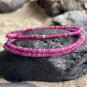 Pode incluir: Uma pulseira enrolada composta por pequenas pedras preciosas rosa-escuras facetadas. A pulseira é enrolada em uma rocha cinza escura. Uma pequena conta prateada é visível na pulseira. O fundo é de cor arenosa.