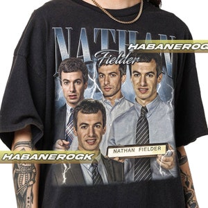 Pode incluir: Camiseta preta com uma estampa gráfica de quatro rostos masculinos. Os homens estão usando ternos e gravatas. O texto "Nathan Fielder" está impresso em uma placa dourada no canto inferior direito do gráfico.