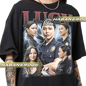 May include: Black t-shirt with a graphic print of a woman in a police uniform. The text "Lucy Chen" is printed on the shirt. The graphic is surrounded by lightning bolts.