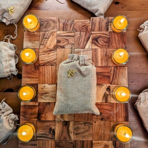 Puede incluir: Una tabla de cortar de madera con un patrón de tablero de ajedrez, rodeada de pequeñas bolsas de arpillera con abalorios de abejas y velas amarillas.