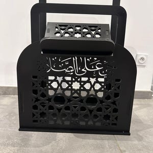 May include: Black wooden Quran stand with intricate laser cut designs and Arabic calligraphy. The stand has a hinged top and a shelf for holding a book.
