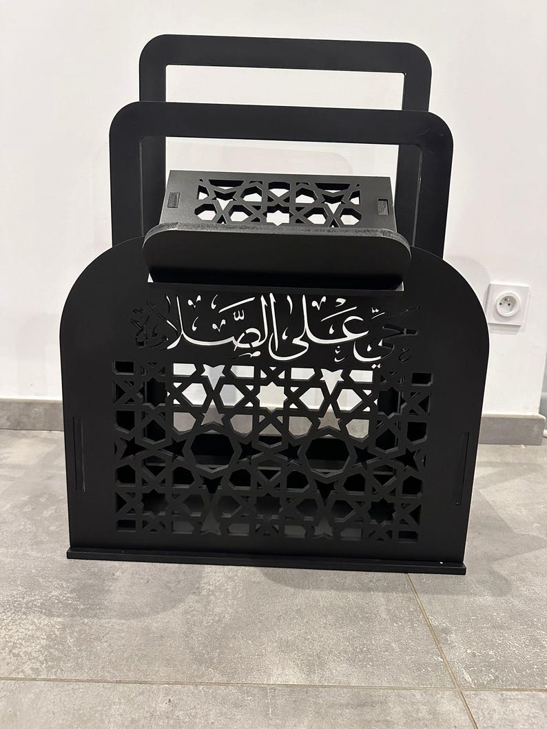 May include: Black wooden Quran stand with intricate laser cut designs and Arabic calligraphy. The stand has a hinged top and a shelf for holding a book.
