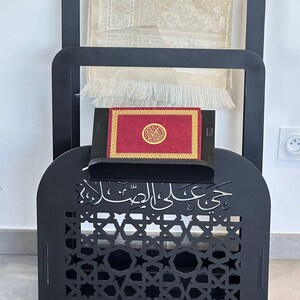 May include: Black wooden prayer stand with intricate cutouts and Arabic calligraphy. It features a shelf for a book and a hook for a prayer rug.