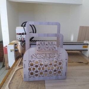 May include: A wooden box with intricate geometric patterns and Arabic calligraphy is being cut out by a CNC machine. The box is made of light brown wood and has a delicate design.