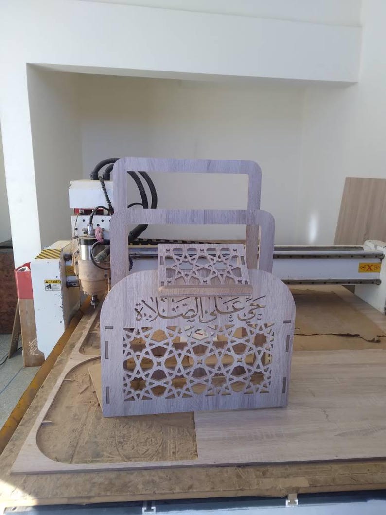 May include: A wooden box with intricate geometric patterns and Arabic calligraphy is being cut out by a CNC machine. The box is made of light brown wood and has a delicate design.
