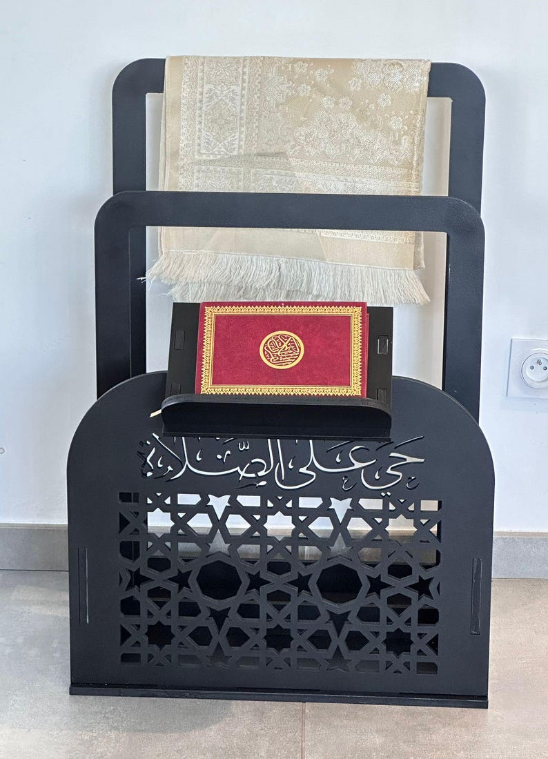 May include: Black wooden prayer stand with intricate cutouts and Arabic calligraphy. It features a shelf for a book and a hook for a prayer rug.