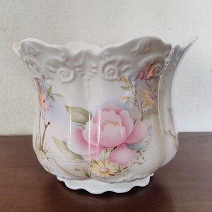 Peut inclure: Un pot de fleurs en céramique blanche avec un bord et une base festonnés, orné d'une grande pivoine rose et d'autres détails floraux. Le pot a une forme bulbeuse et est peint avec des couleurs pastel.