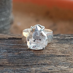 May include: A silver ring featuring a large, clear, faceted quartz stone set in a bezel setting. The stone is pear-shaped and has a rough, natural appearance.
