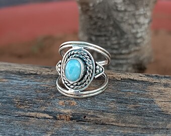 Può includere: Un anello d'argento con una pietra preziosa blu chiaro incastonata in una cornice d'argento decorativa. L'anello ha un design a doppia fascia.