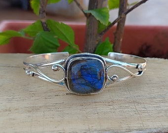 Puede incluir: Un brazalete de plata con una gran piedra de labradorita azul de forma cuadrada. El brazalete tiene un diseño delicado y calado con espirales y pequeñas cuentas.