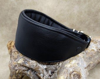 May include: A black leather collar with a wide, padded design. The collar features a buckle closure and a snap button. The item is displayed on a piece of driftwood, showcasing its texture and shape.