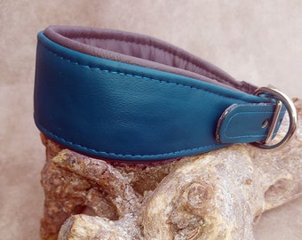 May include: A teal leather dog collar with a padded brown interior and silver-colored hardware. The collar is designed with a curved shape and features teal stitching along the edges. The collar is resting on a piece of wood.