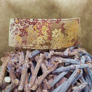 May include: A gold and red patterned fabric collar for a pet. The collar is sitting on a pile of brown sticks.
