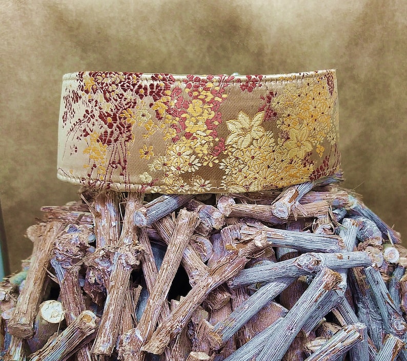 May include: A gold and red patterned fabric collar for a pet. The collar is sitting on a pile of brown sticks.