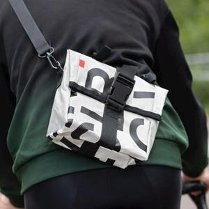 Puede incluir: Un bolso bandolera blanco con tipografía negra llamativa y un pequeño cuadrado rojo. El bolso cuenta con una correa negra ajustable y un cierre de hebilla de plástico negro, llevado sobre la espalda mientras se monta en bicicleta con chaqueta.