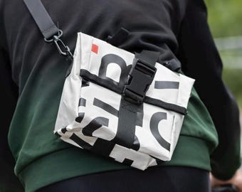 May include: A white crossbody bag with bold black typography and a small red square accent. The bag features a black adjustable strap and a black plastic buckle closure, worn across the back while riding a bicycle in a black and green jacket.