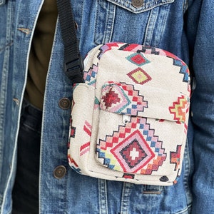 Puede incluir: Un peque&ntilde;o bolso bandolera beige con un estampado geom&eacute;trico colorido. El bolso tiene una correa negra ajustable y un cierre de cremallera.