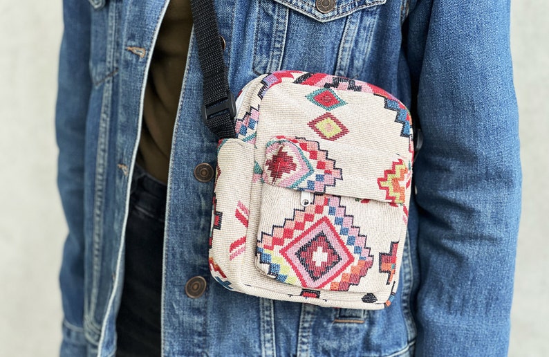 Puede incluir: Un peque&ntilde;o bolso bandolera beige con un estampado geom&eacute;trico colorido. El bolso tiene una correa negra ajustable y un cierre de cremallera.