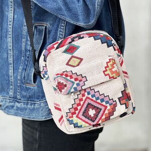 Puede incluir: Un peque&ntilde;o bolso bandolera beige con un estampado geom&eacute;trico colorido. El bolso tiene una correa ajustable negra y un cierre de cremallera.